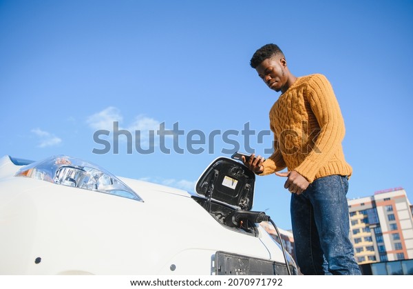 Electric cars, EV concept, eco friendly fuel.\
Portrait of young smiling black man, recharging his modern luxury\
electric car