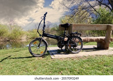 Zündapp Electric Folding Bike Leaning Against A Massive Rustic Wooden Bench In Front Of A River Under Sensible Cloudy Sky In Gifhorn, Germany, April 18, 2020