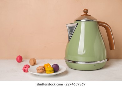 Electric kettle and plate with macaroons on table near brown wall