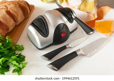 Electric knife sharpener. The plastic body is gray-black. A kitchen knife in the sharpener. In the foreground are kitchen knives. On the back there is bread and greens. Light background.