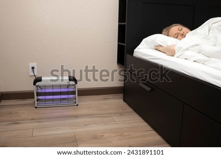 Electric Mosquito or Insect Zapper With Blue Purple Lights Turned on near Sleeping Person in Bed in Bedroom. Bug Killer Lamp on Wooden Floor in Room. Fly Trap Indoor. Horizontal Plane.