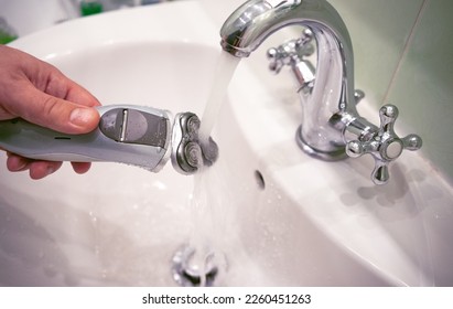 An electric razor in a man's hand. Washing an electric shaver under running water in a faucet. Daily care of shaving trimmer blades.