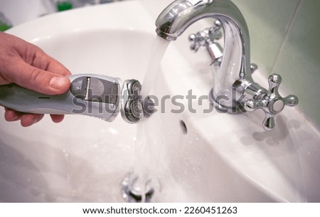 An electric razor in a man's hand. Washing an electric shaver under running water in a faucet. Daily care of shaving trimmer blades.