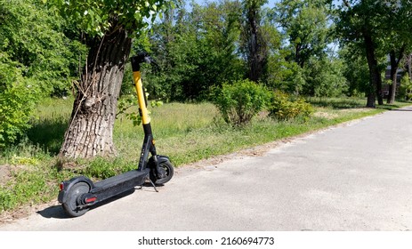 Electric Scooter Or E-scooter Parked Under  Tree On The Path. Alternative Modern Eco-friendly Means Of Transportation In The City For Domestic And Tourists, Visitors. Environmental Care And Urban Life