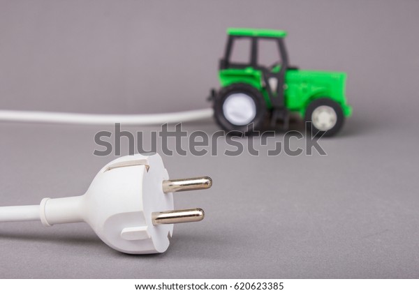 The electric tractor is charged from the\
outlet. Ecotech on the farm. A white plug from the socket is\
connected to a tractor on a gray\
background