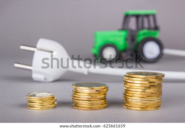 The electric\
tractor is charged from the outlet. Ecotech on the farm. A white\
plug from the socket is connected to a tractor on a gray\
background. A stack of coins,\
savings