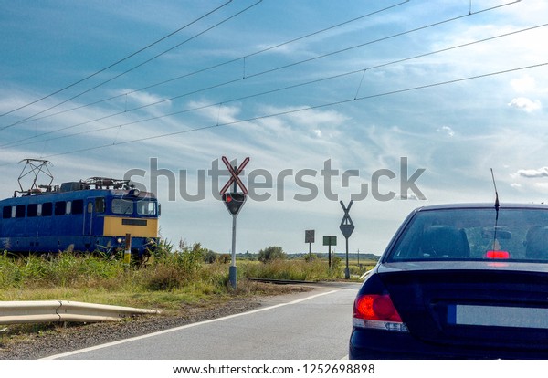 The electric train approaching railroad crossing.\
Car standing in front of the railway crossing with traffic light\
and road sign without a\
barrier