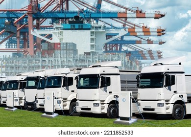 Electric Trucks In The International Seaport Against On A Background Of A Ship Loaded With Containers. 