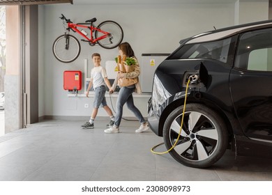 Electric vehicle charging station in private home with happy mother and son walking alongside, blurred family leaving the house, with a bicycle hanging on the wall. Horizontal. - Powered by Shutterstock