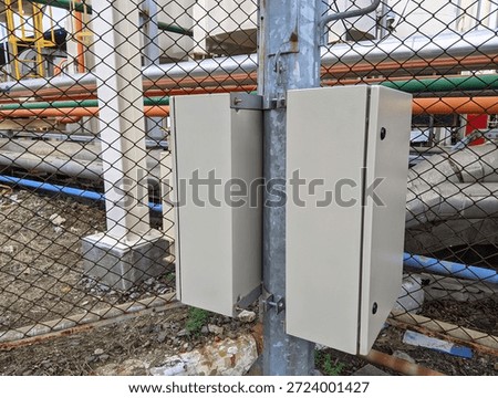 ​Industrial electrical control box mounted on metal post in a chemical plant or factory setting with colorful pipes and chain link fence background.