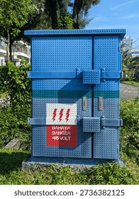 Electrical distribution box painted with a creative LEGO brick pattern design, featuring a red danger warning sign in Indonesian and English 'Bahaya Danger 415 Volt', located outdoors in a green urban area.