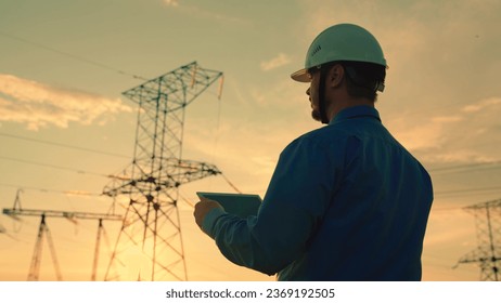 An electrical engineer in hard hat work with digital tablet near power pole. Industry concept. Silhouette electrical engineer with tablet computer, power unit tower with electricity. Sale electricity