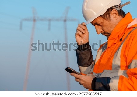Electrical engineer using smartphone out in the field close to electricity pylons, selective focus