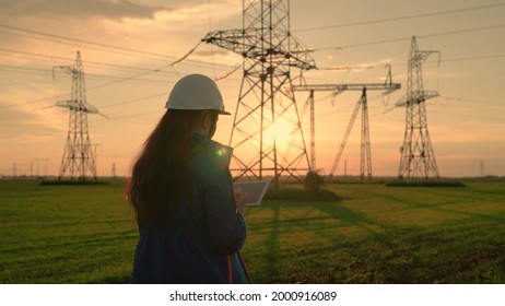 An Electrical Engineer, Woman Working With Computer Tablet, Works With Electricity Next To An Electric Tower. Industry Of Energy Business Technologies. An Electrical Engineer Works On Digital Tablet