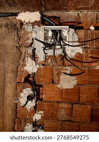 Electrical junction box with cables inside a house under construction. The photo focuses on the junction box with incoming and outgoing electrical wires. Exposed cables, unfinished wiring, and a plastic or metal box mounted on a bare wall. The background shows rough construction materials. Industrial, technical, and realistic view of electrical installation work in progress.