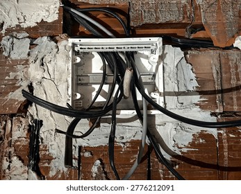 Electrical junction box with cables inside a house under construction. The photo focuses on the junction box with incoming and outgoing electrical wires. Exposed cables, unfinished wiring, and a plastic or metal box mounted on a bare wall. The background shows rough construction materials. Industrial, technical, and realistic view of electrical installation work in progress.