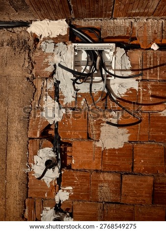Electrical junction box with cables inside a house under construction. The photo focuses on the junction box with incoming and outgoing electrical wires. Exposed cables, unfinished wiring, and a plastic or metal box mounted on a bare wall. The background shows rough construction materials. Industrial, technical, and realistic view of electrical installation work in progress.