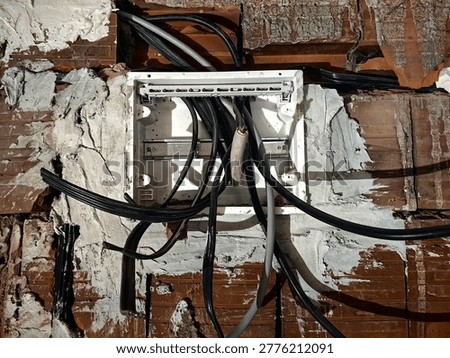 Electrical junction box with cables inside a house under construction. The photo focuses on the junction box with incoming and outgoing electrical wires. Exposed cables, unfinished wiring, and a plastic or metal box mounted on a bare wall. The background shows rough construction materials. Industrial, technical, and realistic view of electrical installation work in progress.