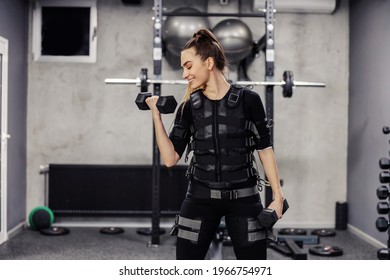 Electrical muscle stimulation, EMS training studio. A sporty girl wearing an EMS suit doing dumbbell arm exercises. A smile and a positive attitude in training. Half of a woman's face with a ponytail