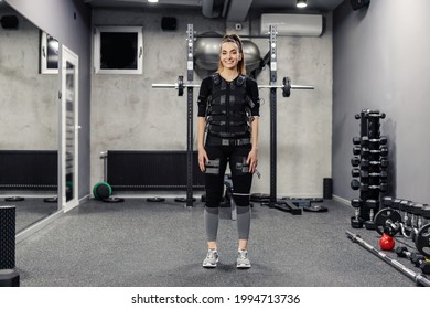 Electrical muscle stimulation. A full-body portrait of a satisfied beautiful woman in a special EMS suit standing in the middle of the gym. Good physical shape, modern concept of training and sports