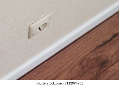 Electrical Outlet On Beige Home Wall With Wallpaper, White Baseboard And Brown Wooden Laminate