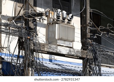 Electrical Pole with Tangled Utility Cables in Urban Area. Electrical pole with tangled utility cables and a transformer near a weathered building facade, showing urban power distribution infrastructure and electrical wiring in a dense city area.