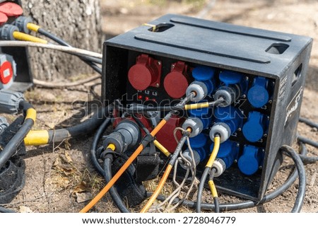 Electrical power distribution box with multiple outlets and cables connected, situated outdoors among trees and natural ground, showcasing a practical setup for outdoor events