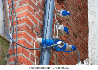 Electrical power sockets and cables attached to a red brick wall with metal conduit pipes, showing outdoor industrial electrical installation details.