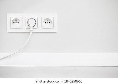Electrical socket isolated on gray wall. White wire plug plugged in. Renovated studio apartment power supply background. Empty copy space triple white plastic power outlet.