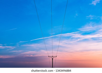 Electrical Substation Utility Pole, State And High Voltage Power Line Silhouette With Blue Cloudy Sky At Sunset. Purple Sunset View Of Electric Tower With Airplane Lines On Blue Sky.