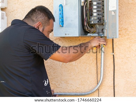An electrical technician attaches metal conduit to a fuse box on the side of a house