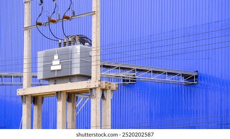 Electrical transformer and high voltage distribution equipment on electric pole with overhead power cable ladder tray next to blue aluminium corrugated metal wall of industrial factory building