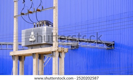 Electrical transformer and high voltage distribution equipment on electric pole with overhead power cable ladder tray next to blue aluminium corrugated metal wall of industrial factory building