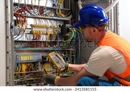 an electrician in a blue helmet, yellow glasses and an orange vest measures electric current with a digital multimeter on a distribution box.