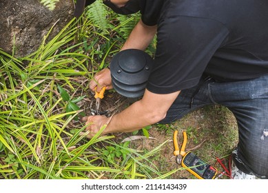 An Electrician Cuts The Faulty Wire Of A Garden Lamp With Pliers. Electrical Repair Service At The Garden Of A House.