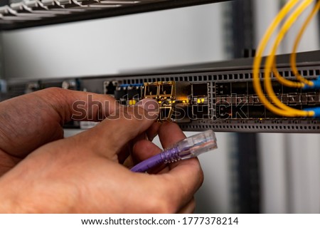 The electrician inserts the SFP module into the router and holds the patch cord. Hands in the frame. Small form-factor pluggable transceiver. Horizontal orientation. 