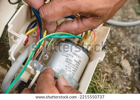 An electrician inspects the wiring connected to CBB65A-1 capacitor inside a window type air conditioner control panel outside.
