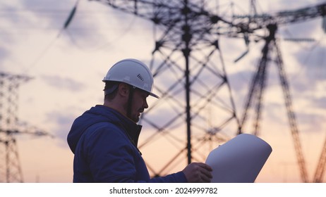 An electrician on the background of high towers of power plants looks at the project for the development of an electrical structure, the expansion of the electrical voltage of volts in the wires