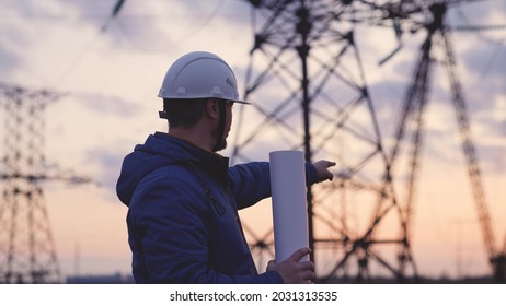 An electrician on the background of high towers of power plants looks at the project for the development of an electrical structure, the expansion of the electrical voltage of volts in the wires
