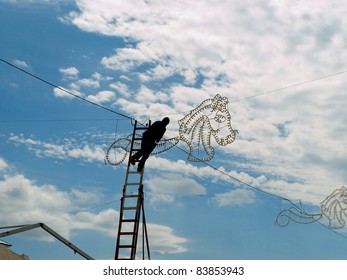 Electrician On A High Ladder Connecting Fair Illuminations , Doing A Dangerous Job At High Altitude