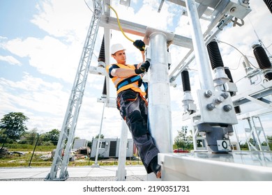 Electrician In Protective Helmet Working On High Voltage Power Lines. Highly Skilled Workmen Servicing The Electricity Grid. Modern Power Station With Power Towers