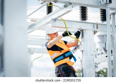 Electrician in protective helmet working on high voltage power lines. Highly skilled workmen servicing the electricity grid. Modern power station with power towers - Shutterstock ID 2190351735