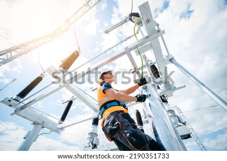 Electrician in protective helmet working on high voltage power lines. Highly skilled workmen servicing the electricity grid. Modern power station with power towers