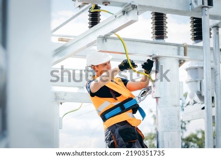 Electrician in protective helmet working on high voltage power lines. Highly skilled workmen servicing the electricity grid. Modern power station with power towers