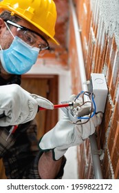 Electrician With A Screwdriver Fixes The Cable In The Switch Of An Electrical System; Wear The Surgical Mask To Prevent The Spread Of Coronavirus. Covid-19, Safety At Work; Construction Industry. 