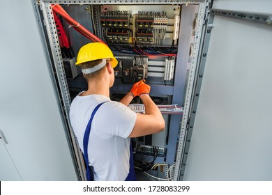 Electrician with screwdriver tighten up switching electric actuator equipment in fuse box. View from the back, a young specialist in a yellow hard hat and overalls. Indoor. - Shutterstock ID 1728083299