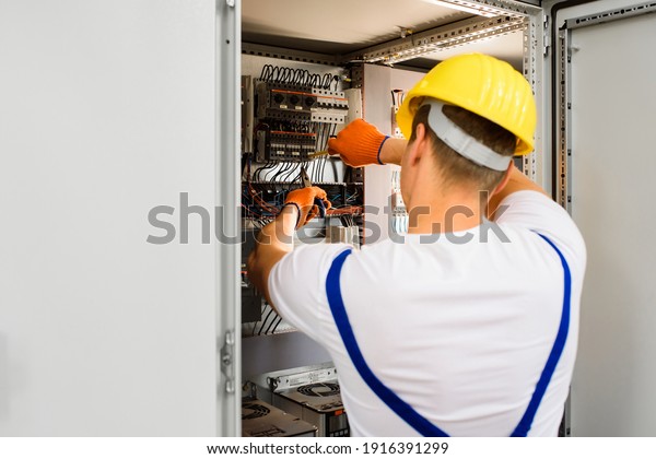Electrician with\
screwdriver tighten up switching electric actuator equipment in\
fuse box. View from the back, a young specialist in a yellow hard\
hat and overalls.\
Indoor.