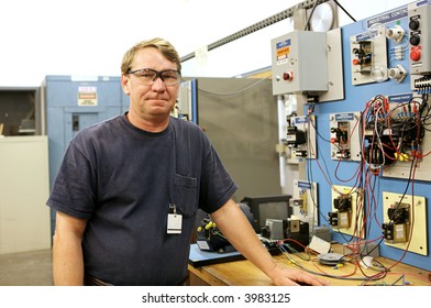 An Electrician In A Technical Education Class Standing Beside A Motor Control Training Board. Actual Electrician With Real Equipment According To National Code And Safety Standards.