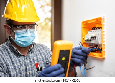 Electrician Worker At Work On An Electrical Panel Protected By Helmet, Safety Goggles And Gloves; Wear The Surgical Mask To Prevent The Spread Of Coronavirus. Construction Industry-Covid 19 Prevention