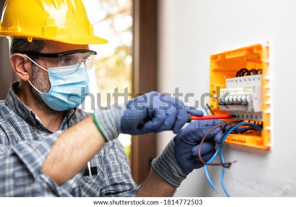 Electrician worker at work on an electrical\
panel protected by helmet, safety goggles and gloves; wear the\
surgical mask to prevent the spread of Coronavirus. Construction\
industry-Covid 19\
Prevention